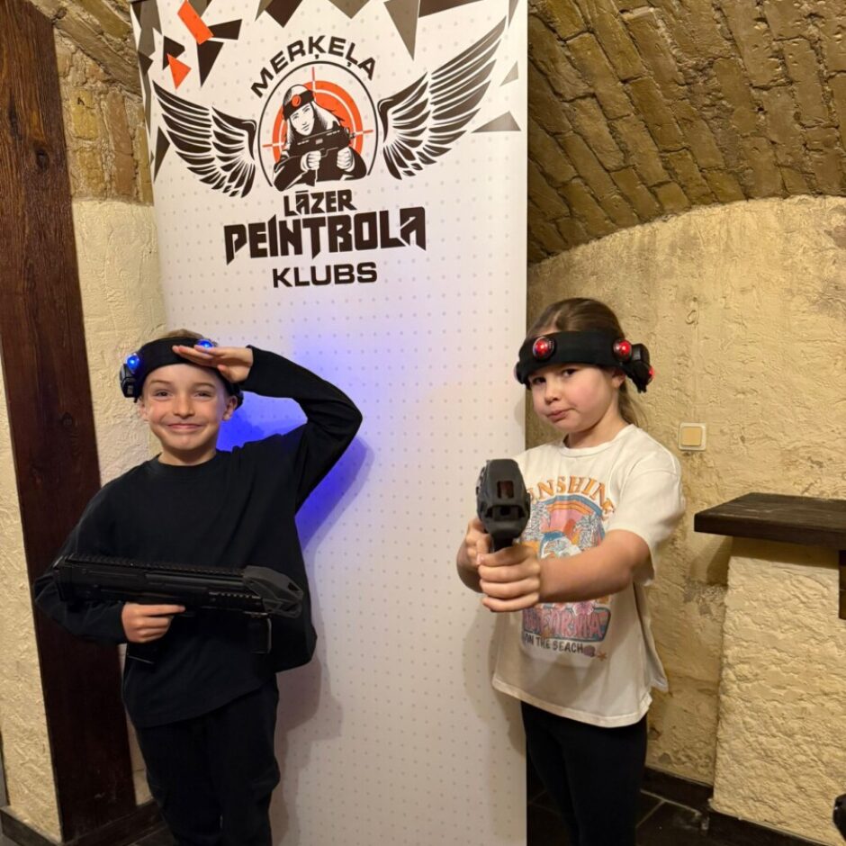 Divi bērni, zēns un meitene, ar lāzertaga aprīkojumu. Zēns kreisajā pusē smaida un salutē, turot lielu lāzeršaujamieroci. Meitene labajā pusē tur mazāku lāzeršaujamieroci. Aiz viņiem ir baneris ar tekstu 