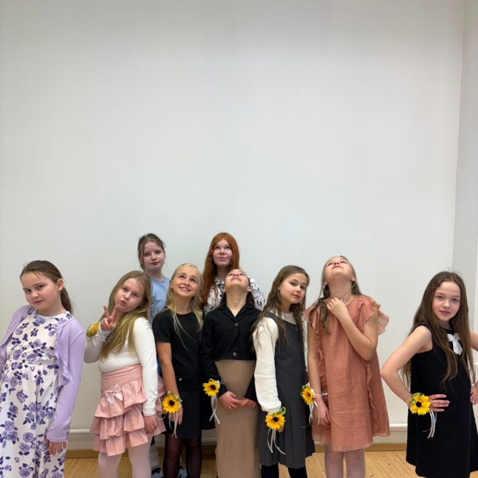 Deviņas jaunas meitenes stāv gaišā telpā ar baltu sienu un koka grīdu, ģērbtas svinīgās kleitās. Daudzām pie rokas ir dzeltenas saulespuķu aproces.