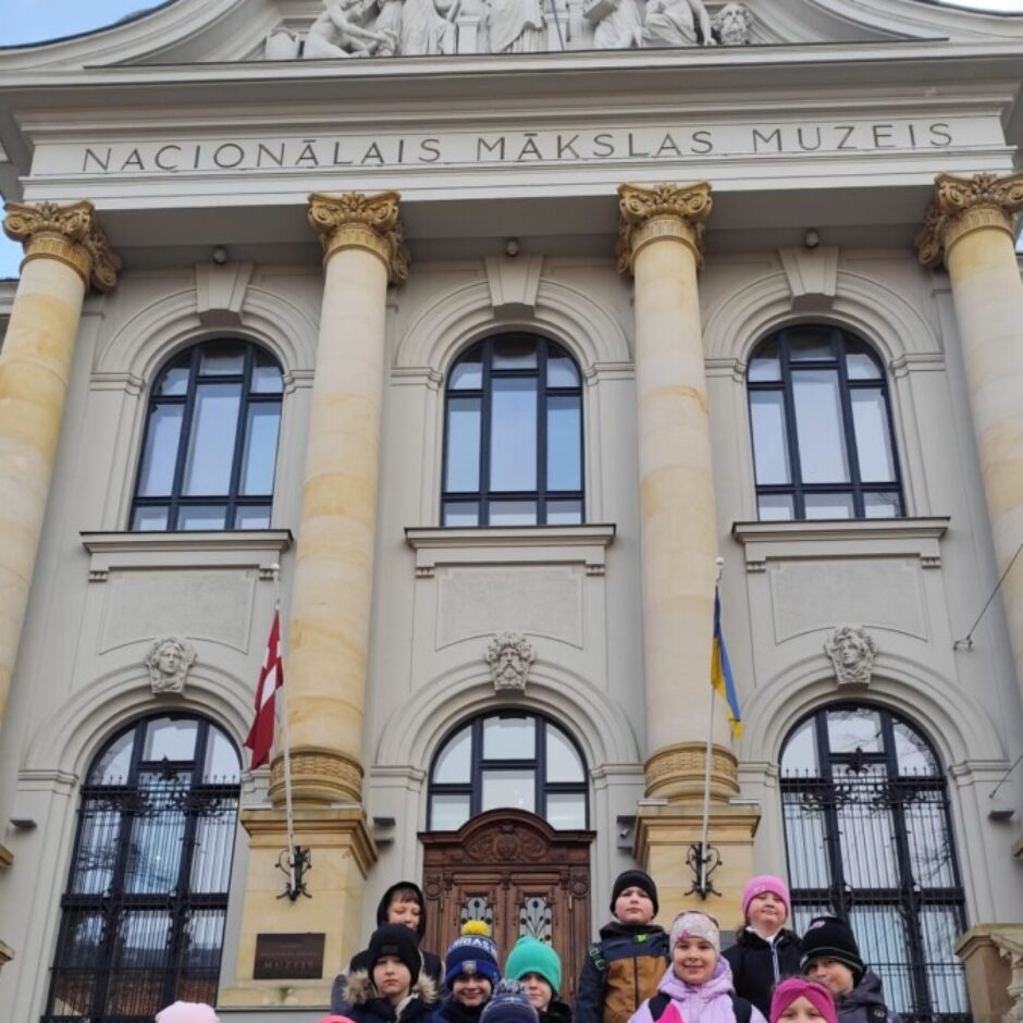 Bērnu grupa stāv uz kāpnēm pie Latvijas Nacionālā mākslas muzeja ēkas ar uzrakstu 