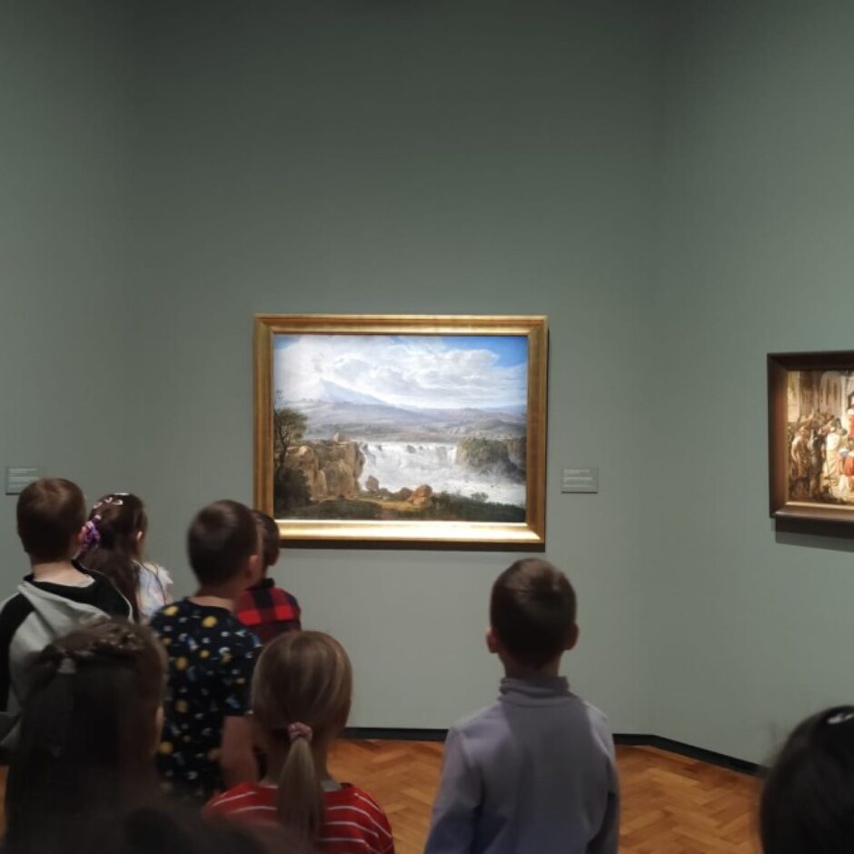 Bērnu grupa un pieaugušais mākslas galerijā aplūko gleznas: vecāka vīrieša portretu, ainavu ar ūdenskritumu un kalniem, un ainu ar daudzām figūrām.