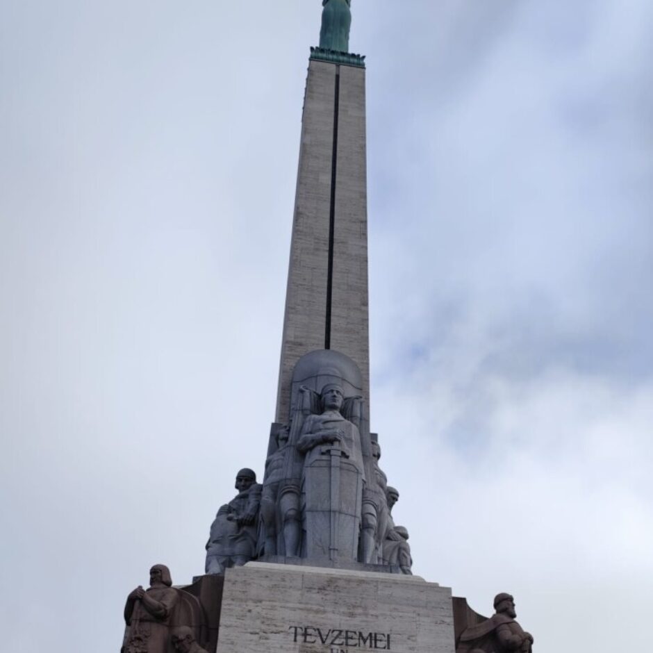 Bērnu grupa stāv pie Brīvības pieminekļa Rīgā, kas ir garš obelisks ar sievietes statuju, kas tur trīs zvaigznes, un skulpturālām grupām pie pamatnes. Uz pieminekļa redzams uzraksts 