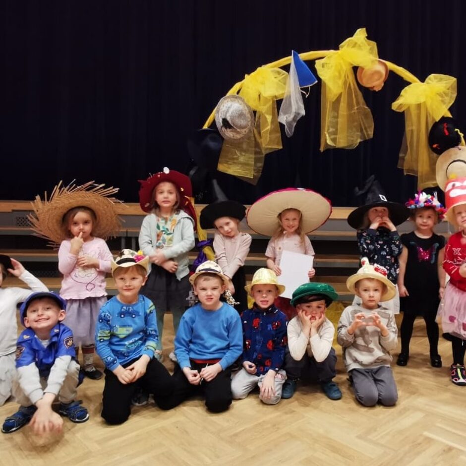 Piecpadsmit bērni, valkājot dažādas iztēles cepures, pozē fotogrāfijai telpās. Viņi stāv un tup uz koka grīdas tumši zila aizkara priekšā. Aizmugurē ir dzeltena arka, kas rotāta ar cepurēm un audumu. Bērnu galvassegas ietver salmu cepures, raganu cepures, platmales un policijas cepuri.