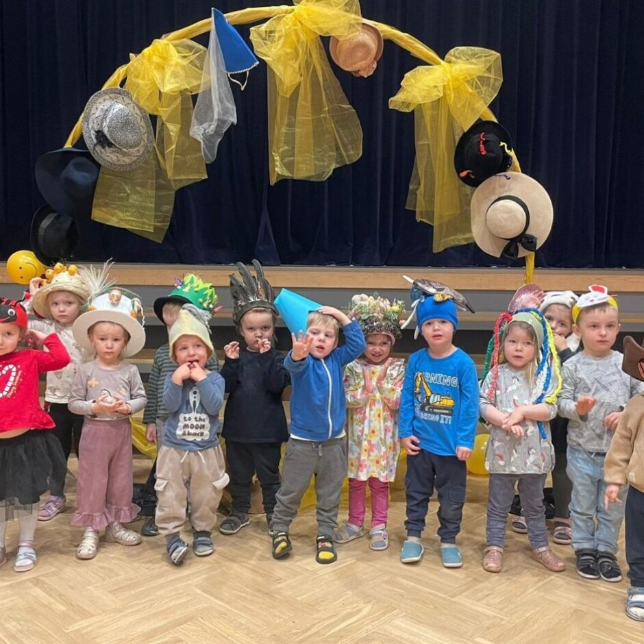Vairāki bērni vecumā no 2 līdz 5 gadiem, tērpušies dažādās cepurēs un kostīmos, stāv rindā uz koka grīdas, skatoties uz priekšu. Starp cepurēm ir vaboļu cepure, kovboju cepure, spalvu cepures un ziedu cepure. Viens zēns valkā zilu kreklu ar uzrakstu 