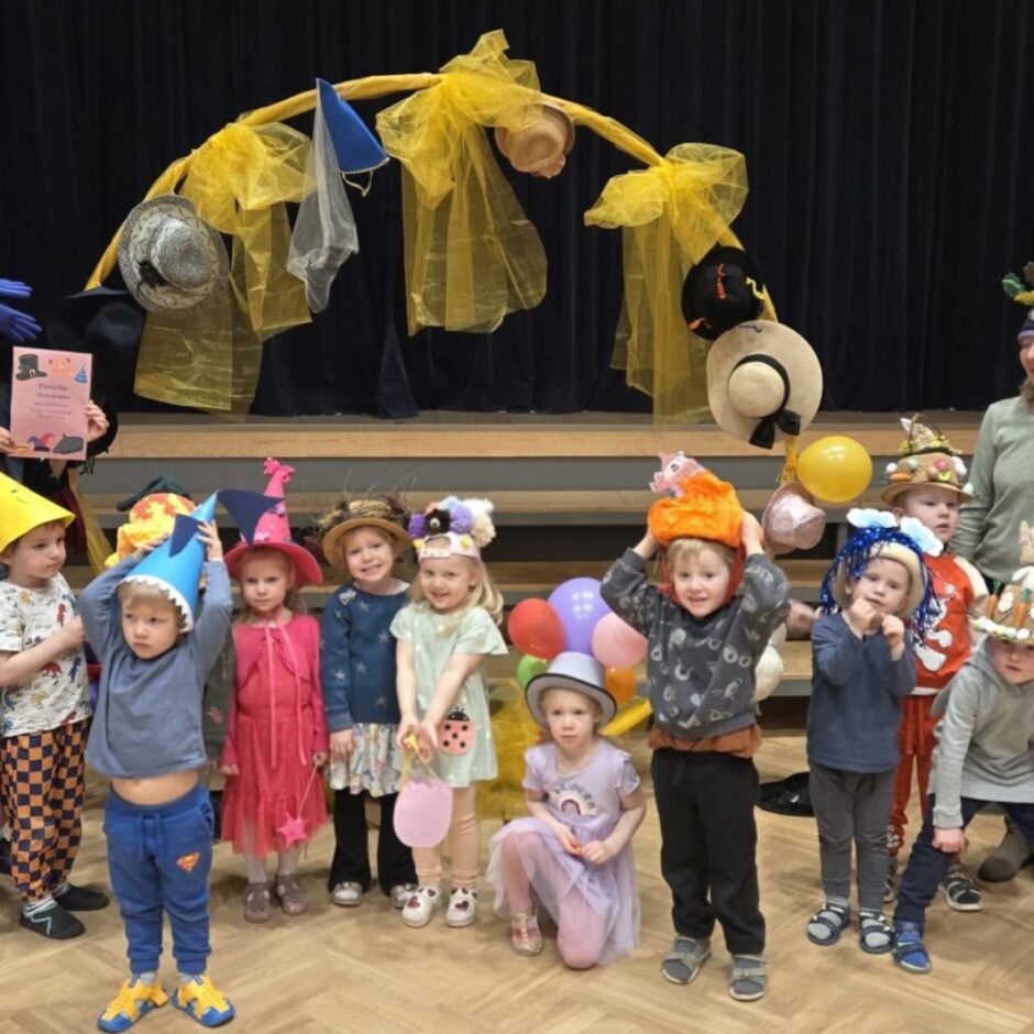 Apmēram piecpadsmit bērni un divas pieaugušas sievietes priecīgi pozē zālē, valkājot dažādas radoši izstrādātas cepures. Sieviete kreisajā pusē, ar lielu zilu cepuri, tur rozā grāmatu, uz kuras rakstīts 