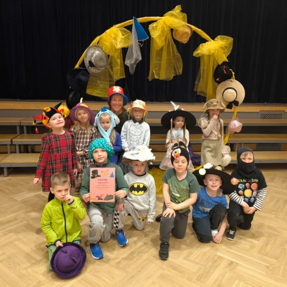 Grupa bērnu un viens pieaugušais, visi ar krāsainām un radošām cepurēm, pozē uz koka grīdas. Aiz viņiem ir skatuve ar dzeltenu arku, kas rotāta ar cepurēm. Viens bērns tur rozā sertifikātu ar tekstu 