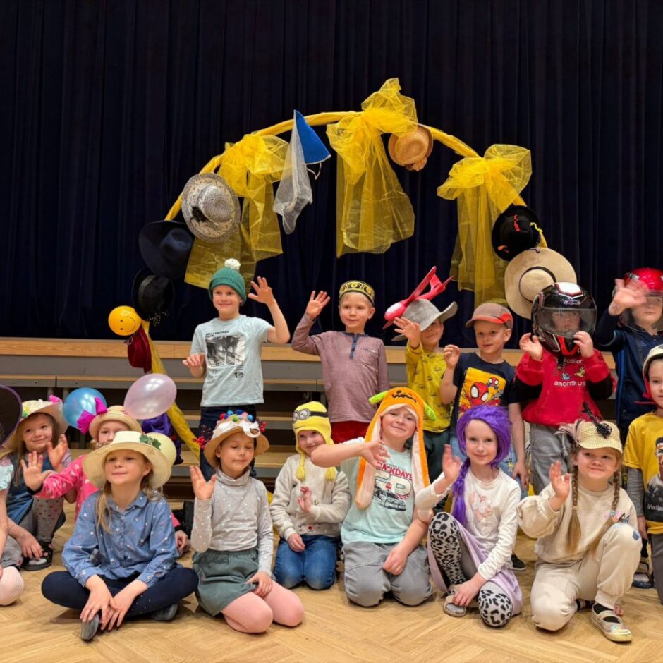 Apmēram 18 smaidoši bērni dažādās cepurēs un kostīmos pozē uz koka grīdas tumši zila aizkara priekšā. Aiz viņiem ir dzeltens dekoratīvs loks, rotāts ar cepurēm. Daudzi bērni māj ar rokām. Starp cepurēm ir raganas cepure, salmu cepures, dzīvnieku cepures, adītas cepures, motocikla un futbola ķiveres. Viena zēna dzeltenajā kreklā redzams uzraksts 