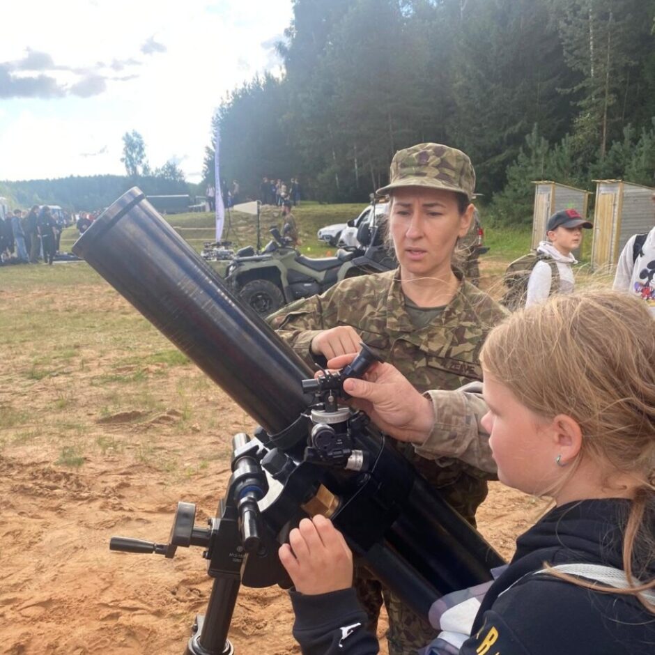 Sieviete militārā formā palīdz jaunai meitenei regulēt lielu melnu optisko tālskati uz statīva. Viņi atrodas ārā, smilšainā apvidū, kur fonā redzami koki un citi cilvēki pasākuma laikā.