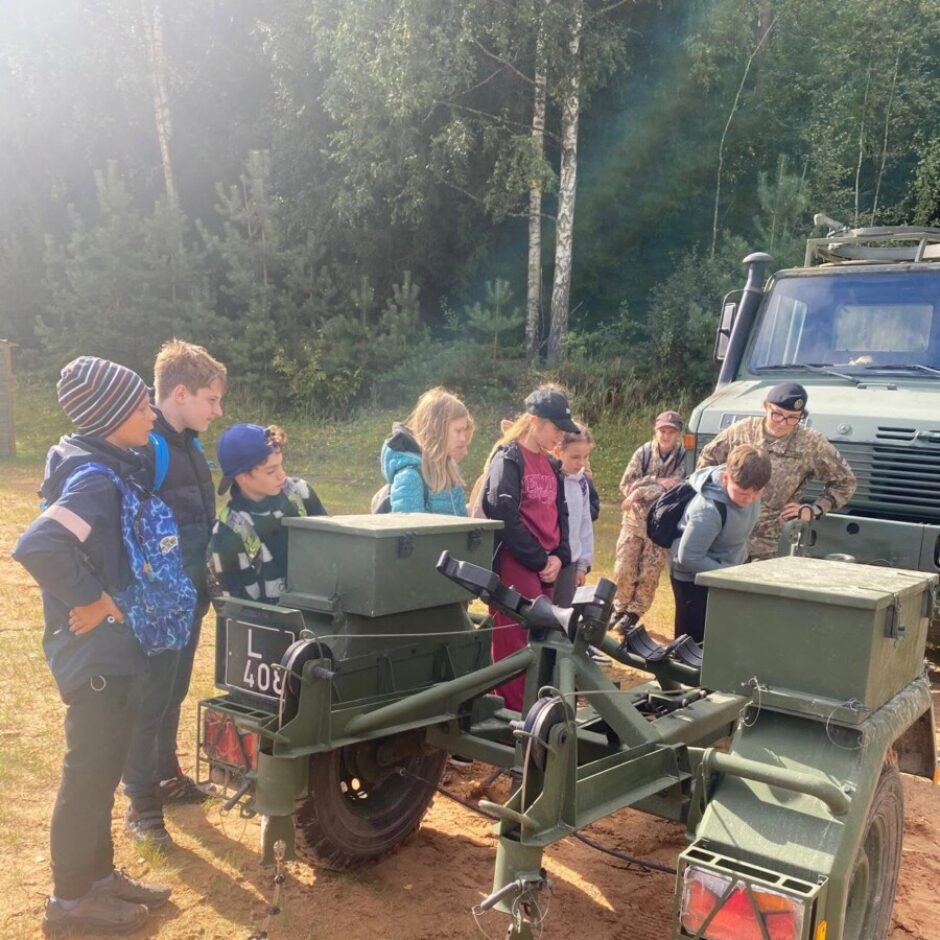 Grupa bērnu kopā ar diviem karavīriem pēta militāru piekabi ar numurzīmi L 407 smilšainā apvidū. Fona redzams militārs transportlīdzeklis un koki.