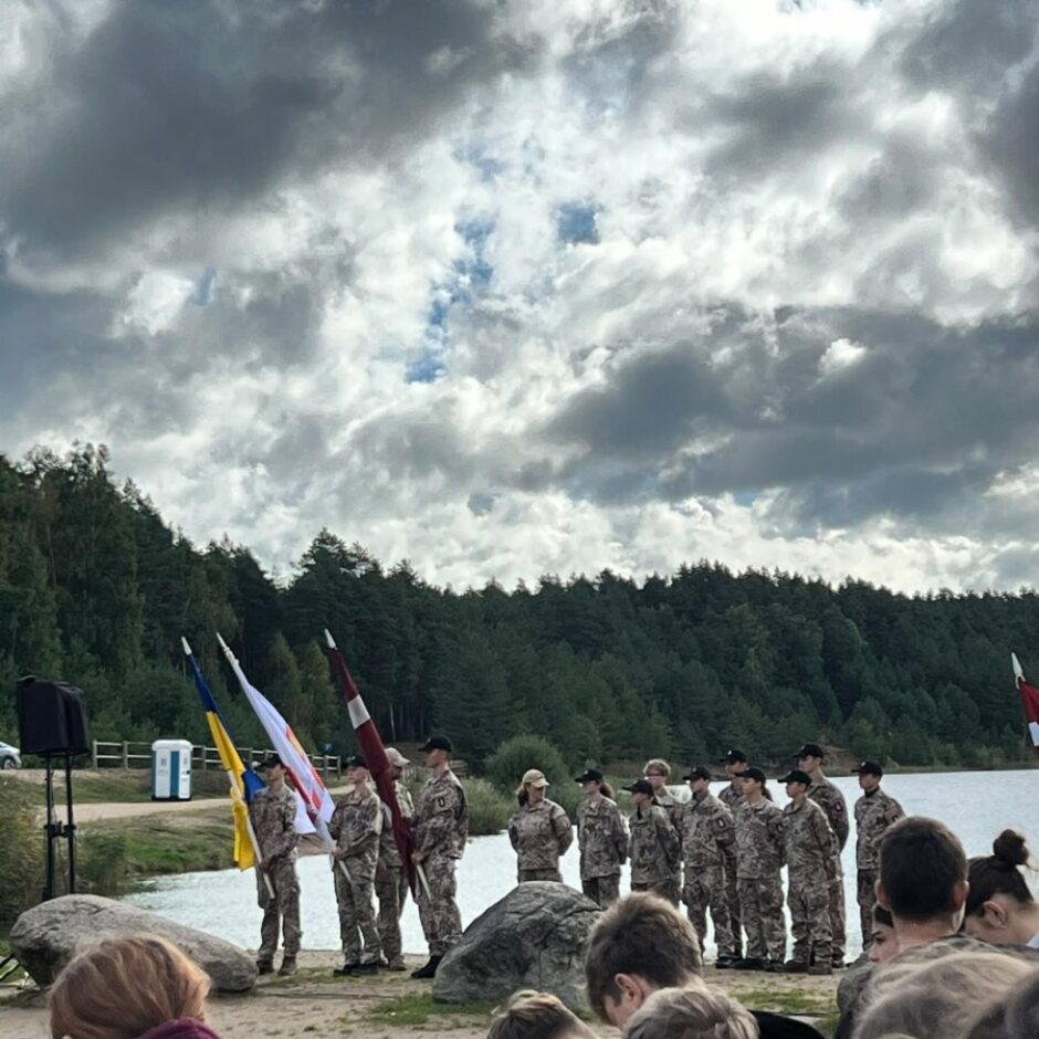 Jaunieši maskēšanās formās stāv ierindā pie ezera krasta, turot Ukrainas, Latvijas un citu karogu. Pasākumu vēro skatītāji priekšplānā, fonā redzams mežs un mākoņainas debesis.