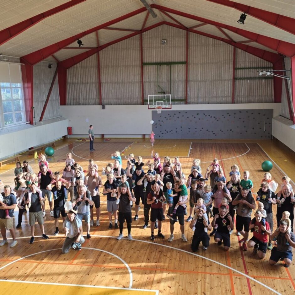 Liela grupa pieaugušo un bērnu stāv sporta zālē, daudzi bērni sēž pieaugušajiem uz pleciem, pozējot kopīgai fotogrāfijai. Fonā redzams basketbola grozs un kāpšanas siena.