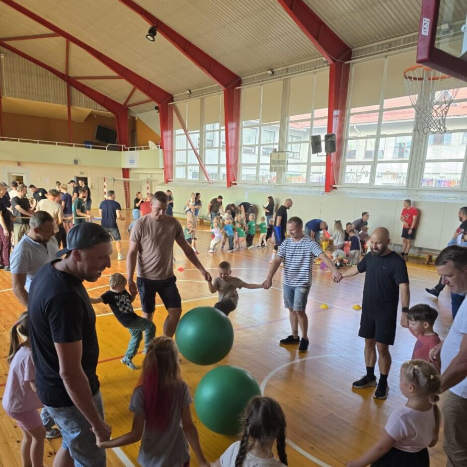 Liela pieaugušo un bērnu grupa gaišā sporta zālē sadevušies rokās veido lokus un līnijas grupu aktivitātē. Uz koka grīdas ir divas lielas zaļas vingrošanas bumbas.