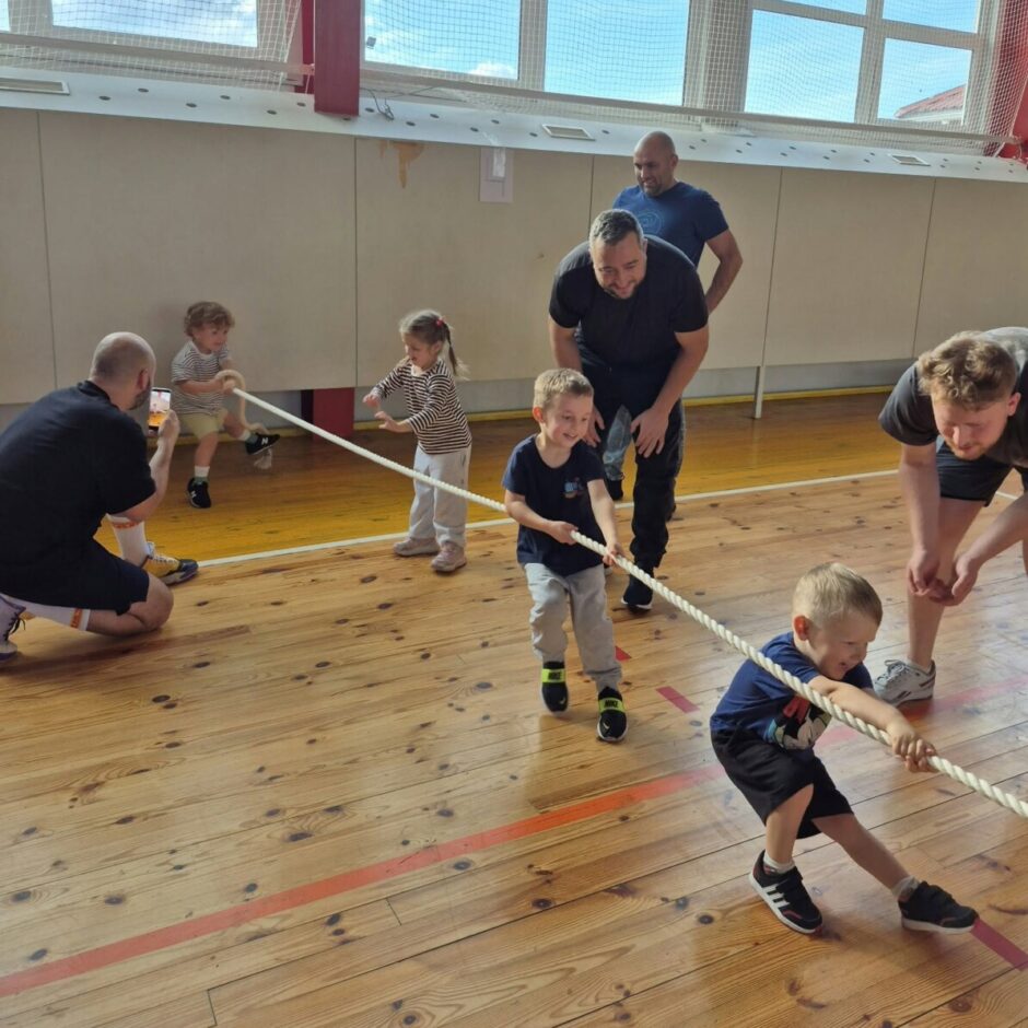 Vairāki bērni un pieaugušie piedalās virves vilkšanā sporta zālē. Divi zēni priekšplānā smaida, stiepjot virvi, savukārt fonā citi bērni un pieaugušie vēro vai piedalās spēlē uz koka grīdas.