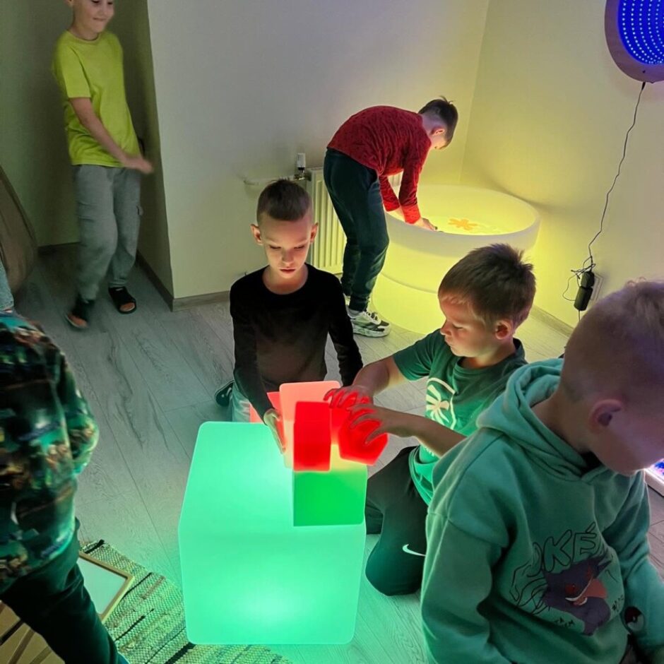 Vairāki zēni spēlējas vāji apgaismotā istabā ar spīdošiem gaismas elementiem. Divi zēni centrā mijiedarbojas ar zaļiem un sarkaniem gaismas klučiem uz liela zaļa spīdoša kuba. Viens zēns priekšplānā sēž uz tirkīzzila šūpojoša spilvena. Fonā zēns noliecas pār lielu, apaļu, spīdošu baltu galdu, kamēr cits zēns stāv blakus.