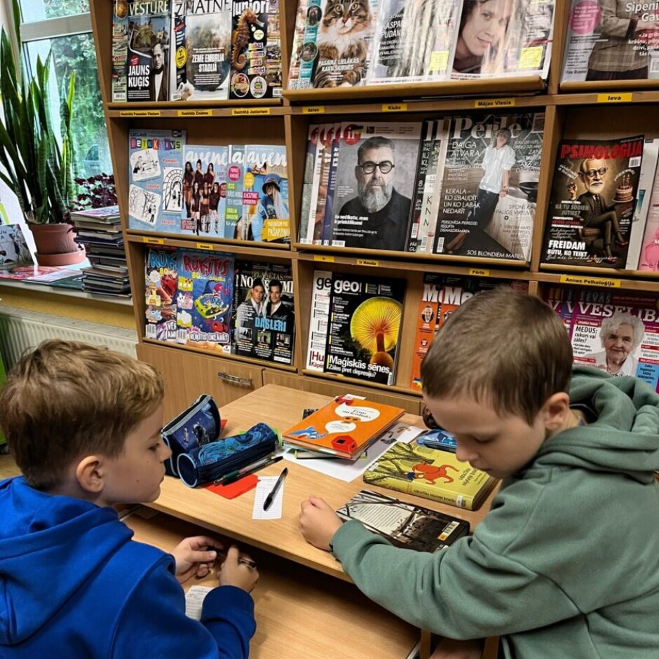 Divi zēni sēž pie koka galda bibliotēkā vai lasītavā. Zilā džemperī tērpts zēns kreisajā pusē raksta, bet zēns zaļā džemperī labajā pusē lasa atvērtu grāmatu. Aizmugurē redzami plaukti ar žurnāliem, tostarp 