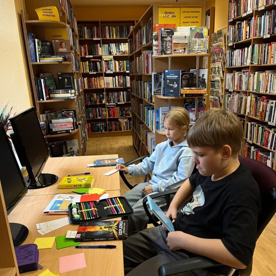 Divi bērni sēž pie galda grāmatu ieskautā bibliotēkā. Meitene skatās datora ekrānā, kamēr zēns tur rokās planšetdatoru. Fonā redzami plaukti ar grāmatām un zīmēm 