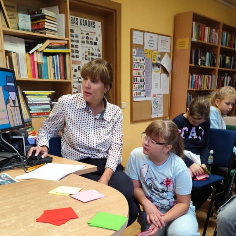 Pieauguša sieviete un bērni bibliotēkā piedalās aktivitātē. Sieviete sēž pie galda, strādā pie datora ar detektīva ilustrāciju ekrānā un skatās uz meiteni ar brillēm, kura tup līdzās galdam, turot rokā sarkanu lapiņu. Uz galda ir kaudzītes ar sarkanām, dzeltenām, rozā un zaļām papīra lapiņām. Fonā grāmatu plaukti un plakāts ar uzrakstu 
