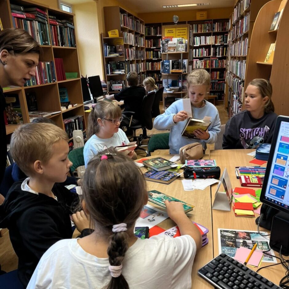 Bērni lasa grāmatas pie galda bibliotēkā, pieaugušais stāv blakus. Fonā redzami grāmatu plaukti un citi bērni, kas strādā pie datoriem. Uz vienas meitenes džempera ir uzraksts 