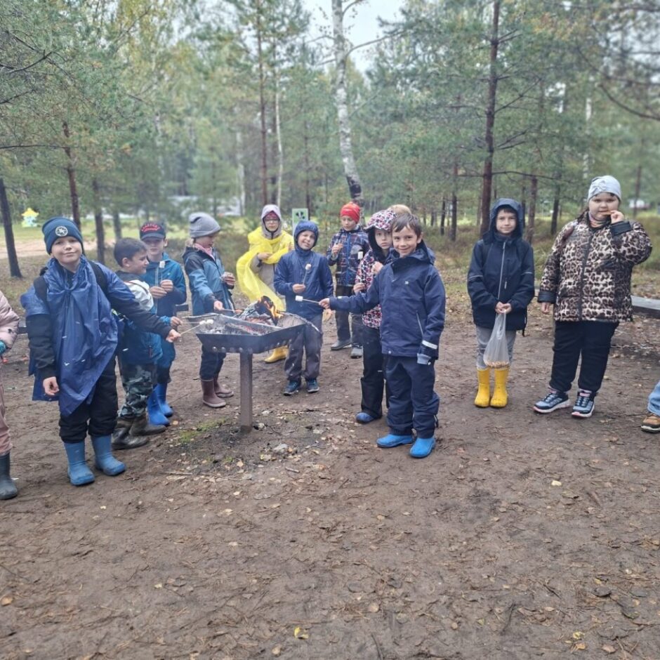 Bērnu grupa mežā cep zefīrus uz grila, valkājot siltas drēbes un zābakus.