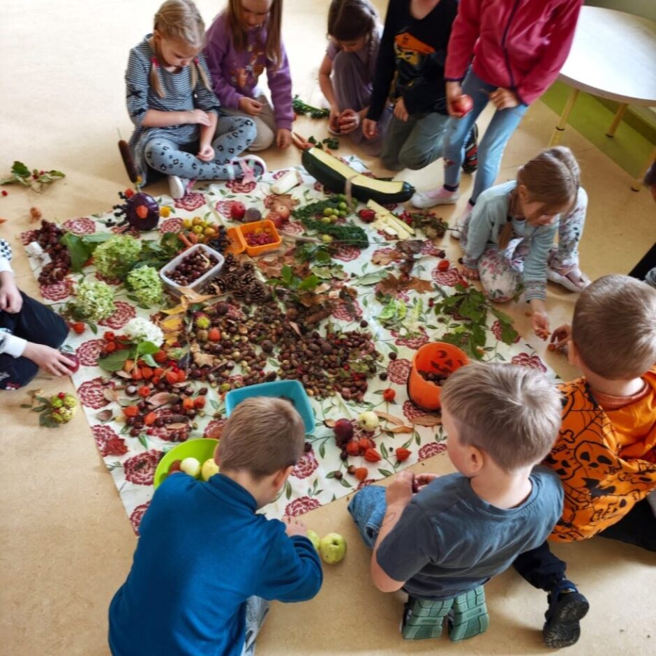 Grupa bērnu sēž uz grīdas ap segu, uz kuras izklāti dažādi rudens dabas materiāli – zīles, kastaņi, lapas, ogas un dārzeņi. Viņi iesaistīti radošā nodarbībā.