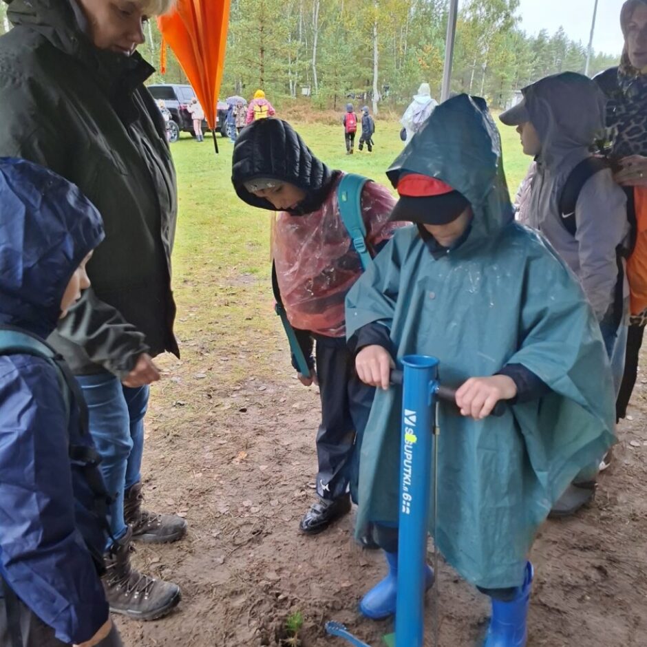 Zēns zaļā pončo un zilajos zābakos stāda koka stādu dubļainā zemē, izmantojot zilu stādīšanas rīku. Apkārt ir citi bērni un pieaugušie lietusmēteļos un ar lietussargu, vērojot stādīšanu meža malā lietainā dienā.