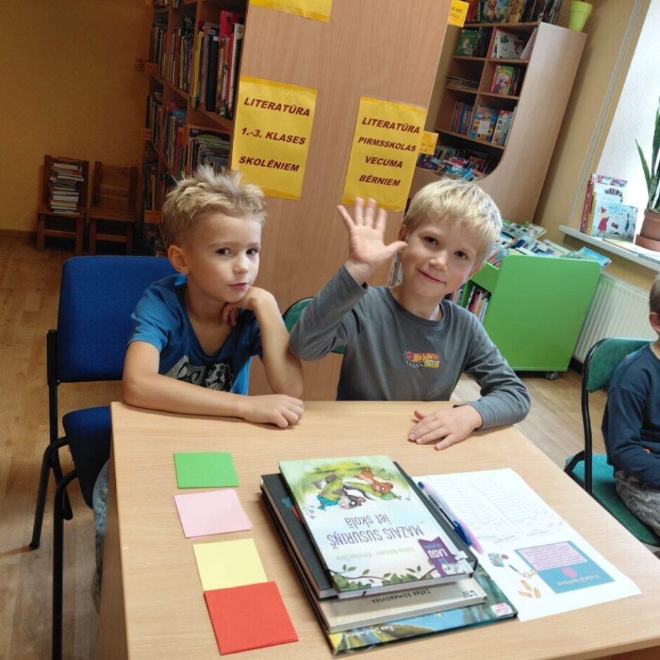 Trīs zēni sēž pie galda bibliotēkā. Kreisais zēns zilā kreklā skatās uz priekšu, atbalstot zodu uz rokas. Vidējais zēns pelēkā kreklā smaida un māj ar roku. Labais zēns tumšā kreklā skatās uz labo pusi. Uz galda ir grāmatas, tostarp viena ar nosaukumu 