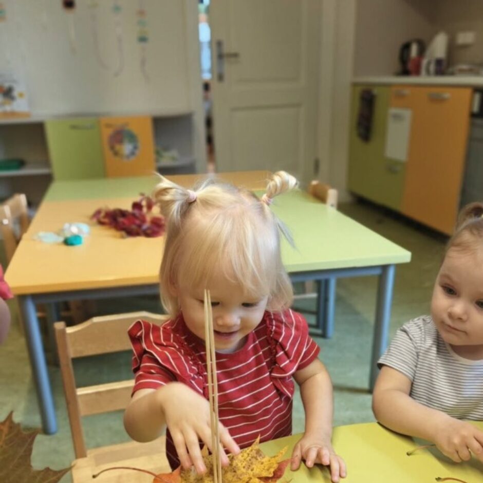 Maza meitene ar gaišām matu copītēm sarkanā svītrainā kreklā smaida, veidojot rudens kompozīciju. Viņa iedur kociņus dzeltenās kļavu lapās, kas novietotas uz zaļa sūnu pušķa baltā traukā. Uz dzeltenzaļa galda izkārtotas dzeltenas un zaļas lapas. Fonā redzams cits bērns un bērnudārza telpas.