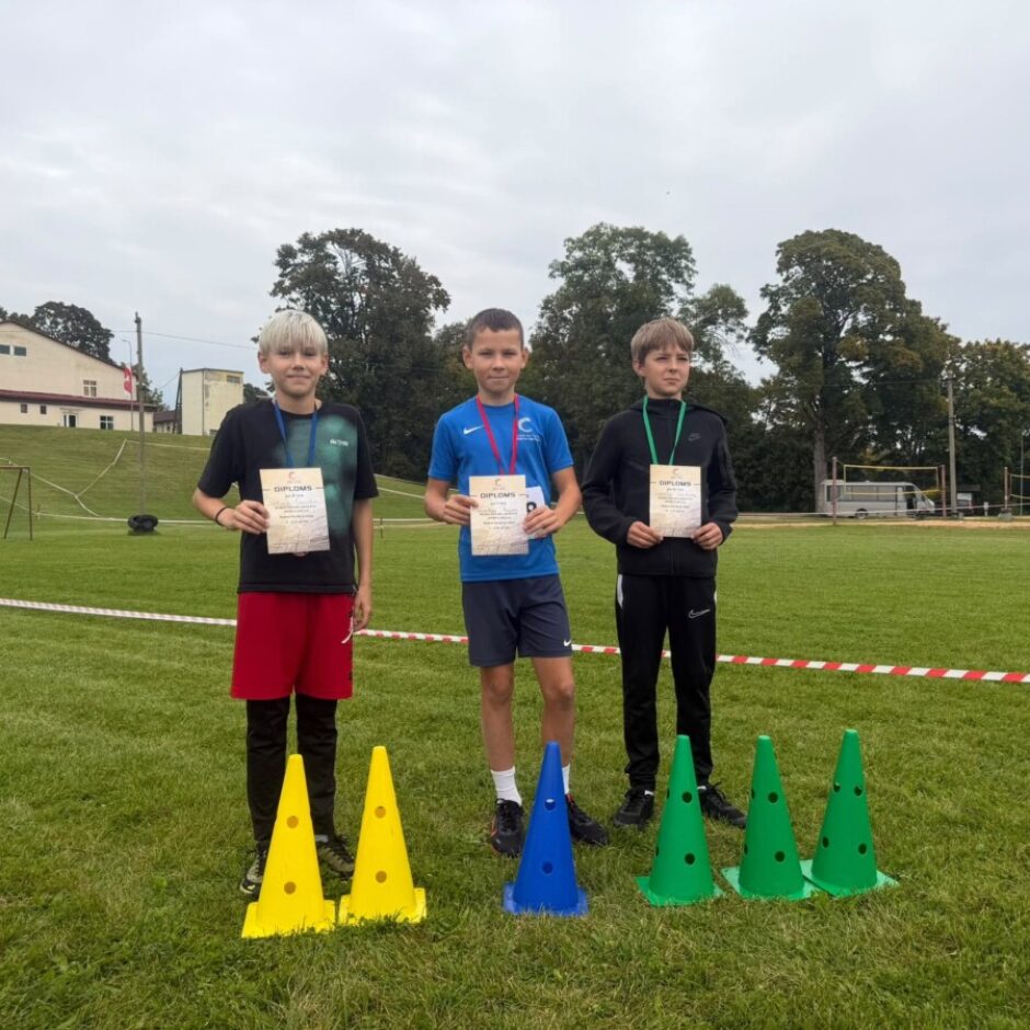 Trīs zēni stāv sporta laukumā ar diplomiem, uz kuriem rakstīts 