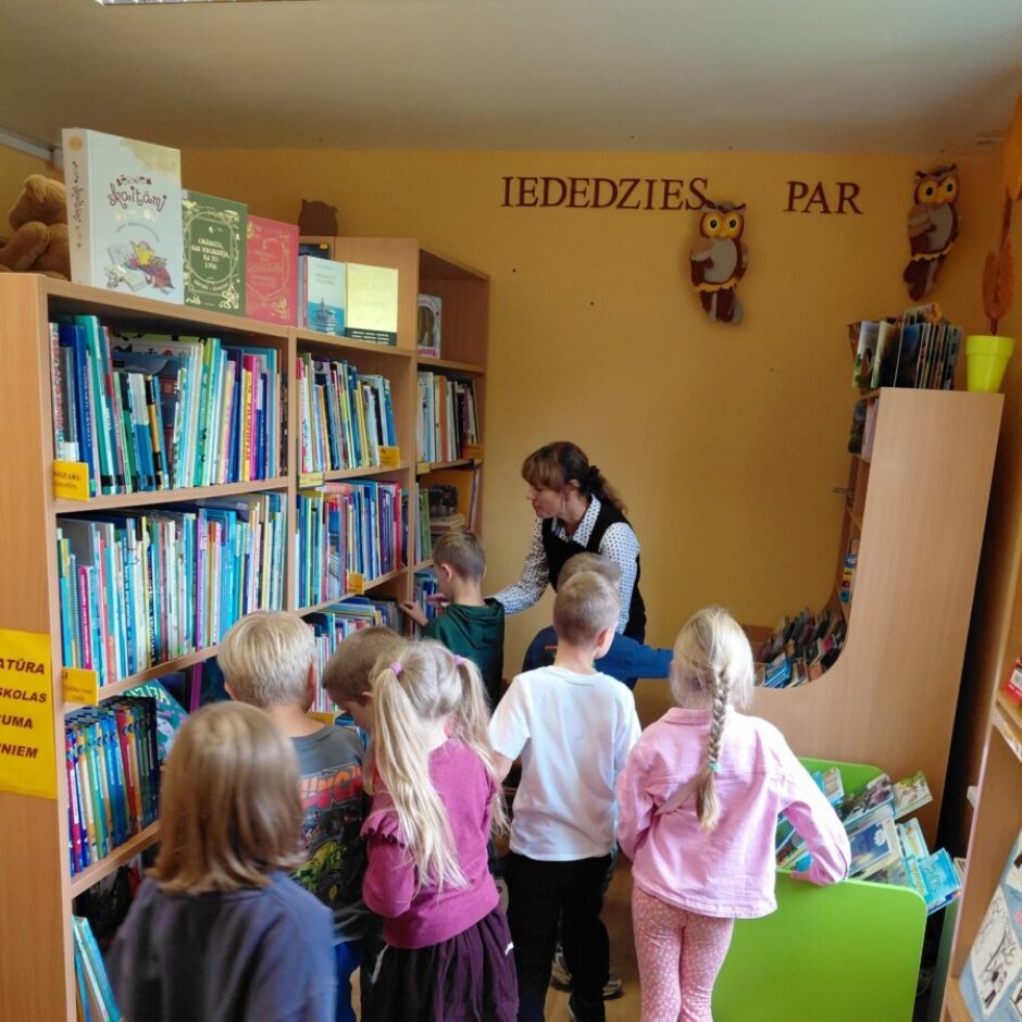 Pieauguša sieviete bērnu bibliotēkas telpā palīdz vairākiem bērniem izvēlēties grāmatas no koka plauktiem. Bērni, kas stāv apkārt, rūpīgi aplūko grāmatas. Uz dzeltenās sienas virs plauktiem ir dekoratīvas pūces.
