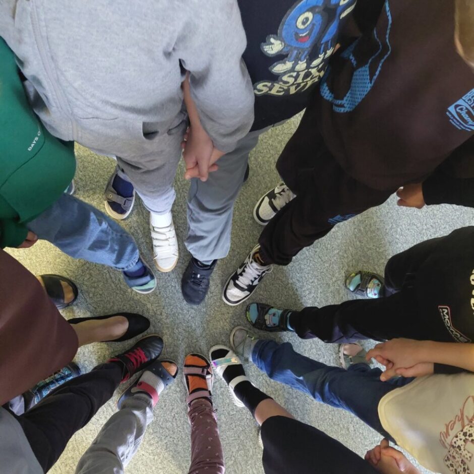 Bērnu grupa, kas turas rokās un stāv aplī, redzama no augšas. Attēlā redzamas viņu kājas un pēdas ar dažādiem apaviem un zeķēm.