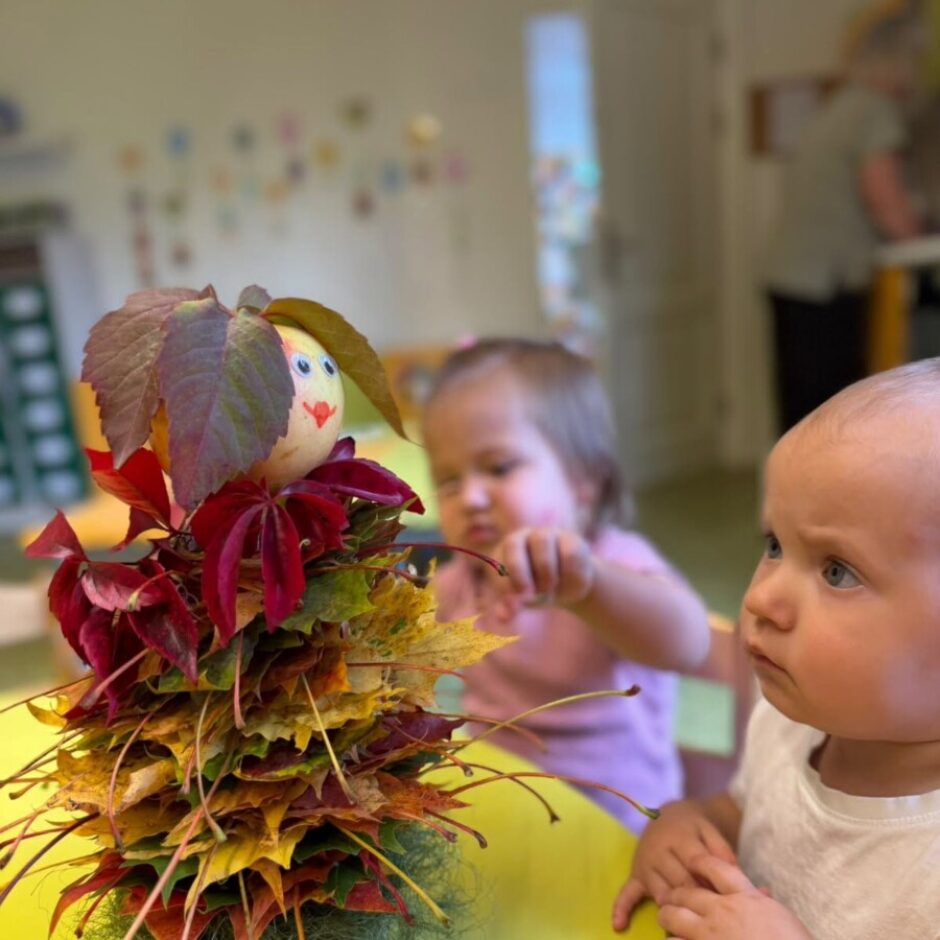 Roka tur krāsainu rudens lapu lelli ar uzzīmētu sejiņu un lapu matiem. Pie dzeltena galda bērnudārzā divi mazi bērni ar interesi skatās uz to.