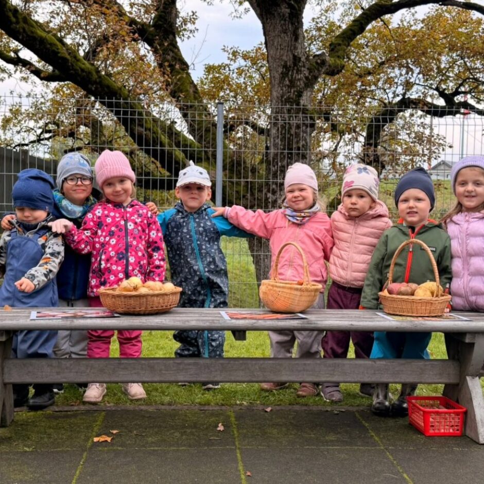 Astoņi mazi bērni rudens apģērbā stāv ārā aiz koka galda, skatoties uz priekšu. Uz galda atrodas trīs pīti grozi ar rudens veltēm – āboliem, kartupeļiem un citiem dārzeņiem, kā arī zīmējumi. Fonā redzams liels koks ar dzeltenīgām lapām.