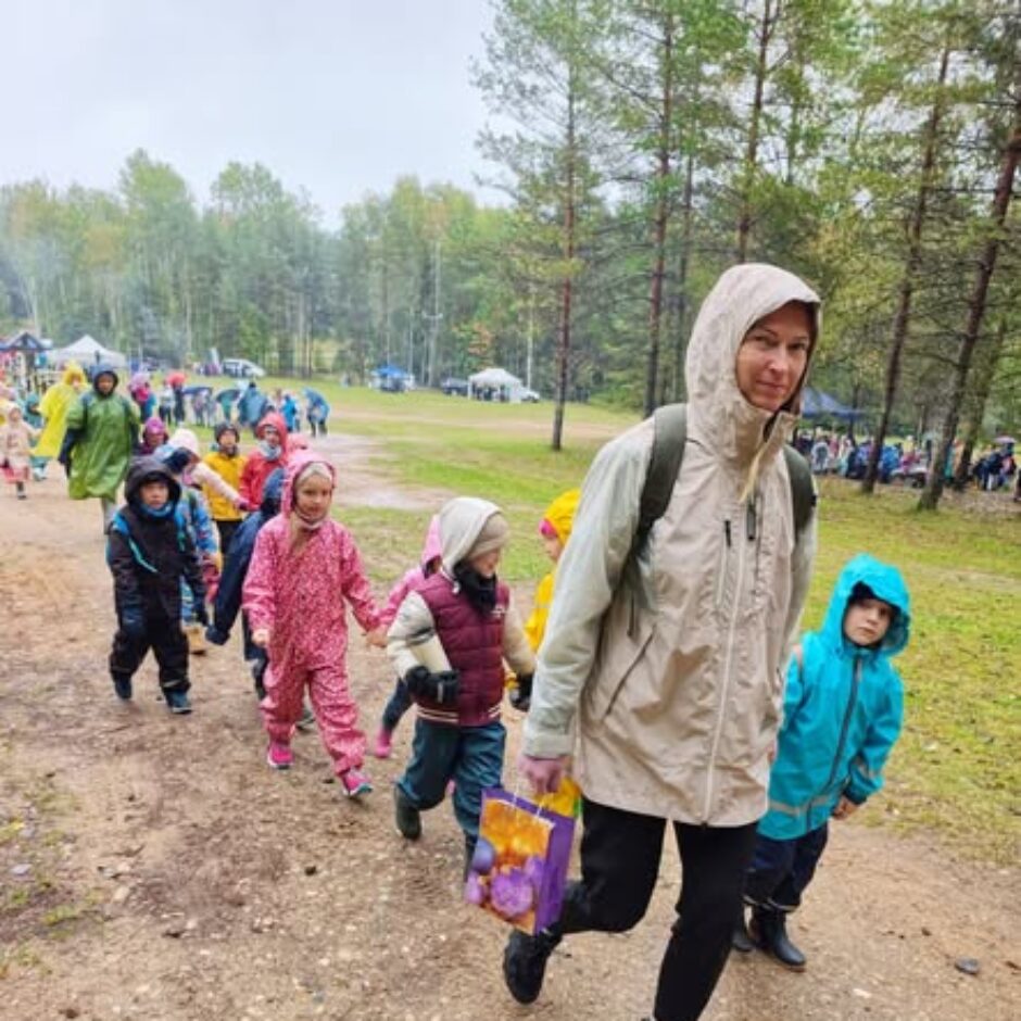 Sieviete gaišā lietusmētelī, turot dāvanu maisiņu, ved bērnu grupu lietus apģērbā pa zemes taku meža malā. Fonā redzami citi pasākuma dalībnieki un teltis lietainā dienā.